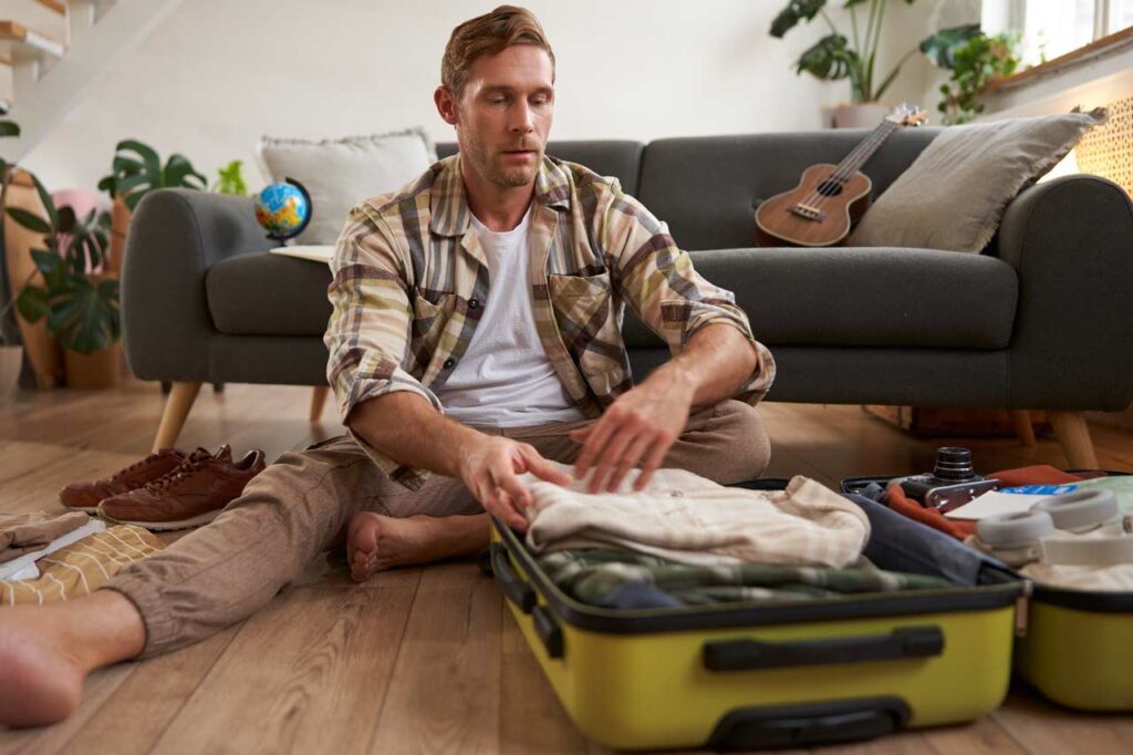 Image of young man, traveller, sitting on floor with opened suitcase, packing clothes for a trip